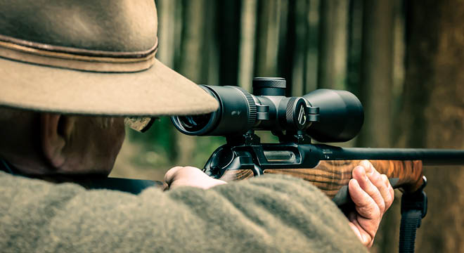 Man, in a wooded setting, looking down his riflescope at his target 